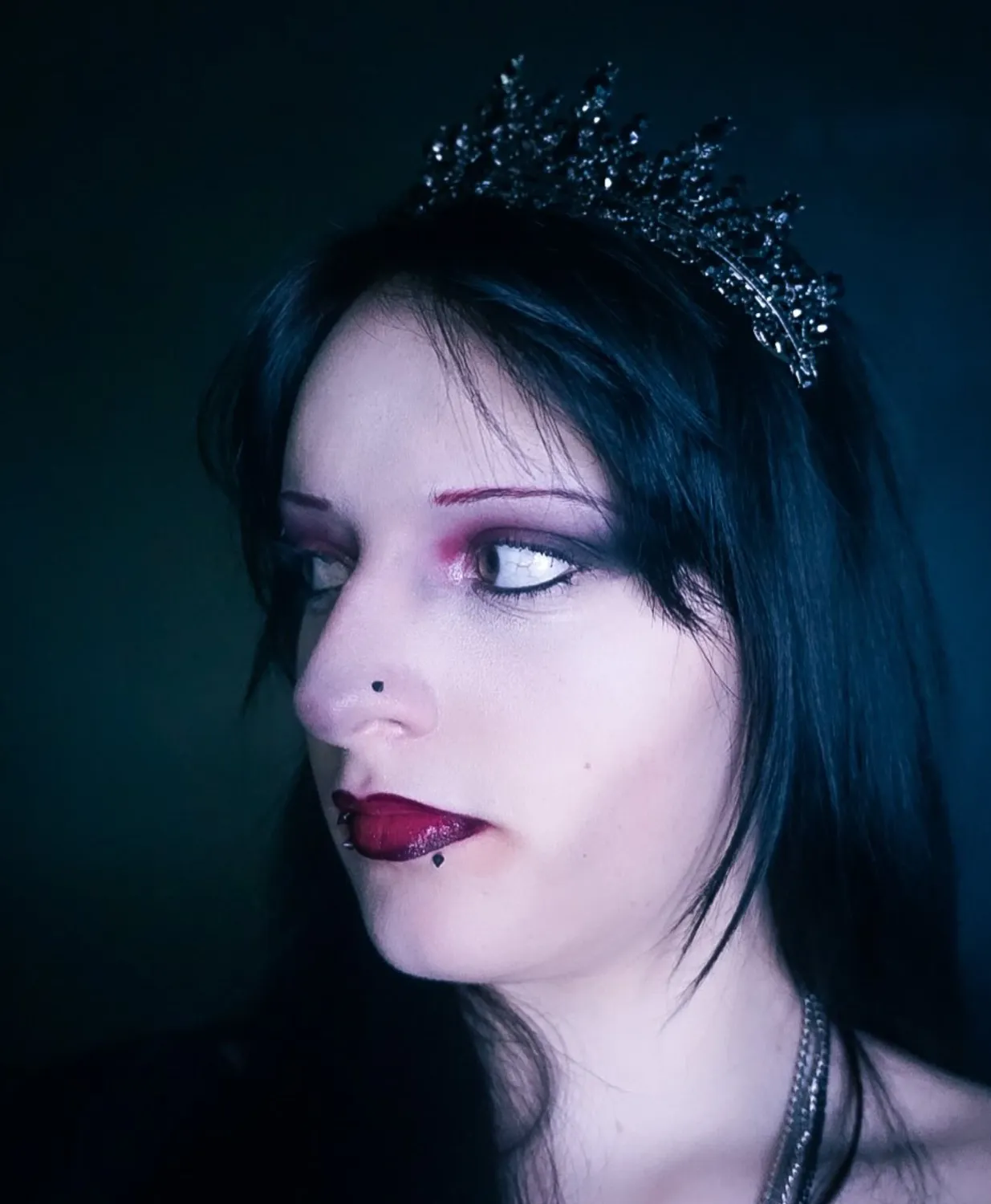 A dark-haired figure in a black crown lit in cool teal, looking off into shadow