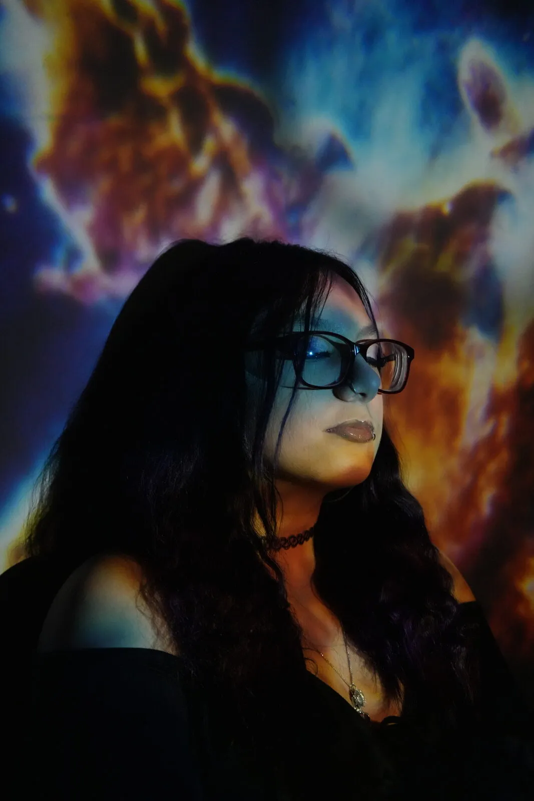 A woman in glasses gazing off camera in front of a swirling projected nebula of orange and blue