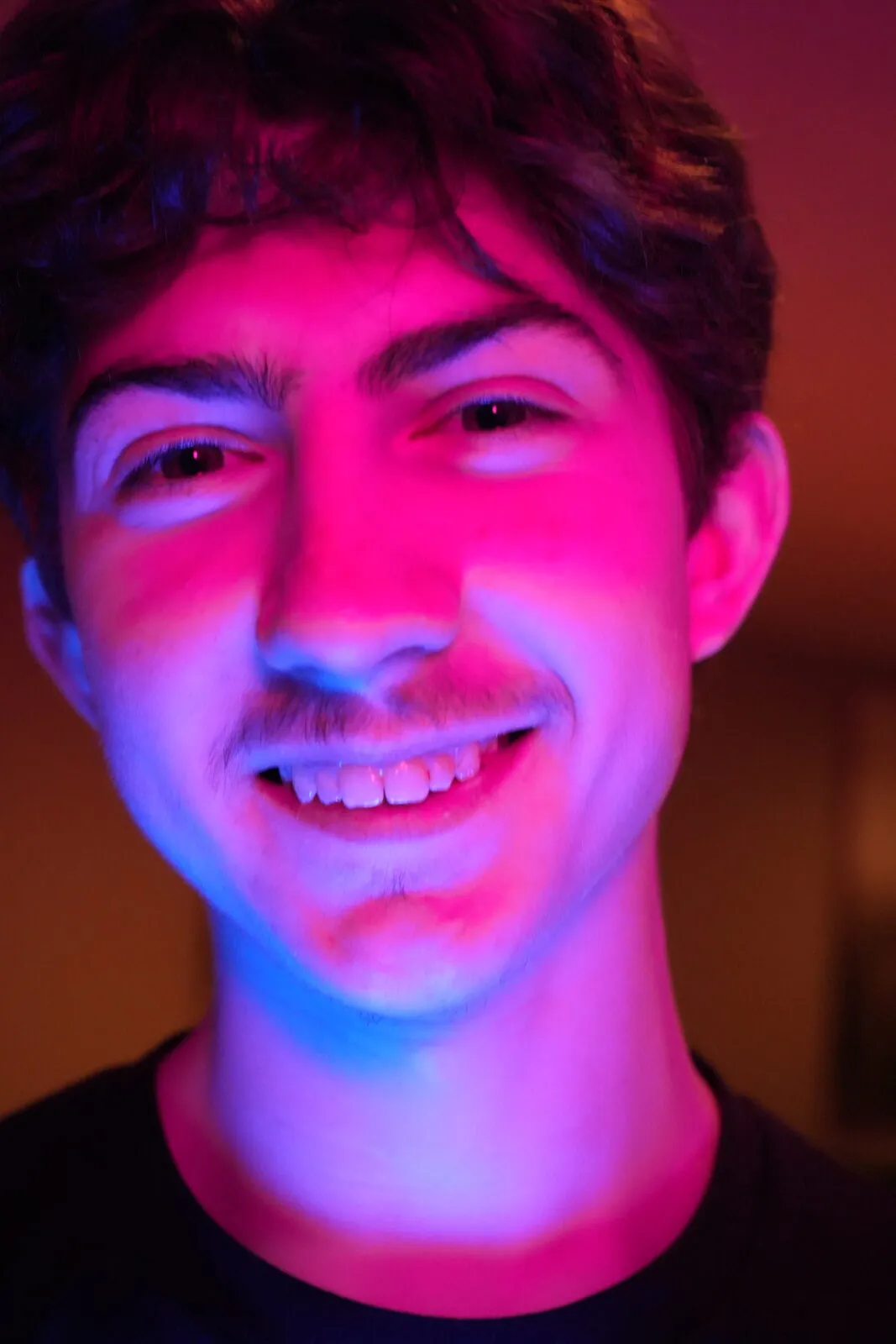 A close headshot of a young man smiling in split pink and blue neon light