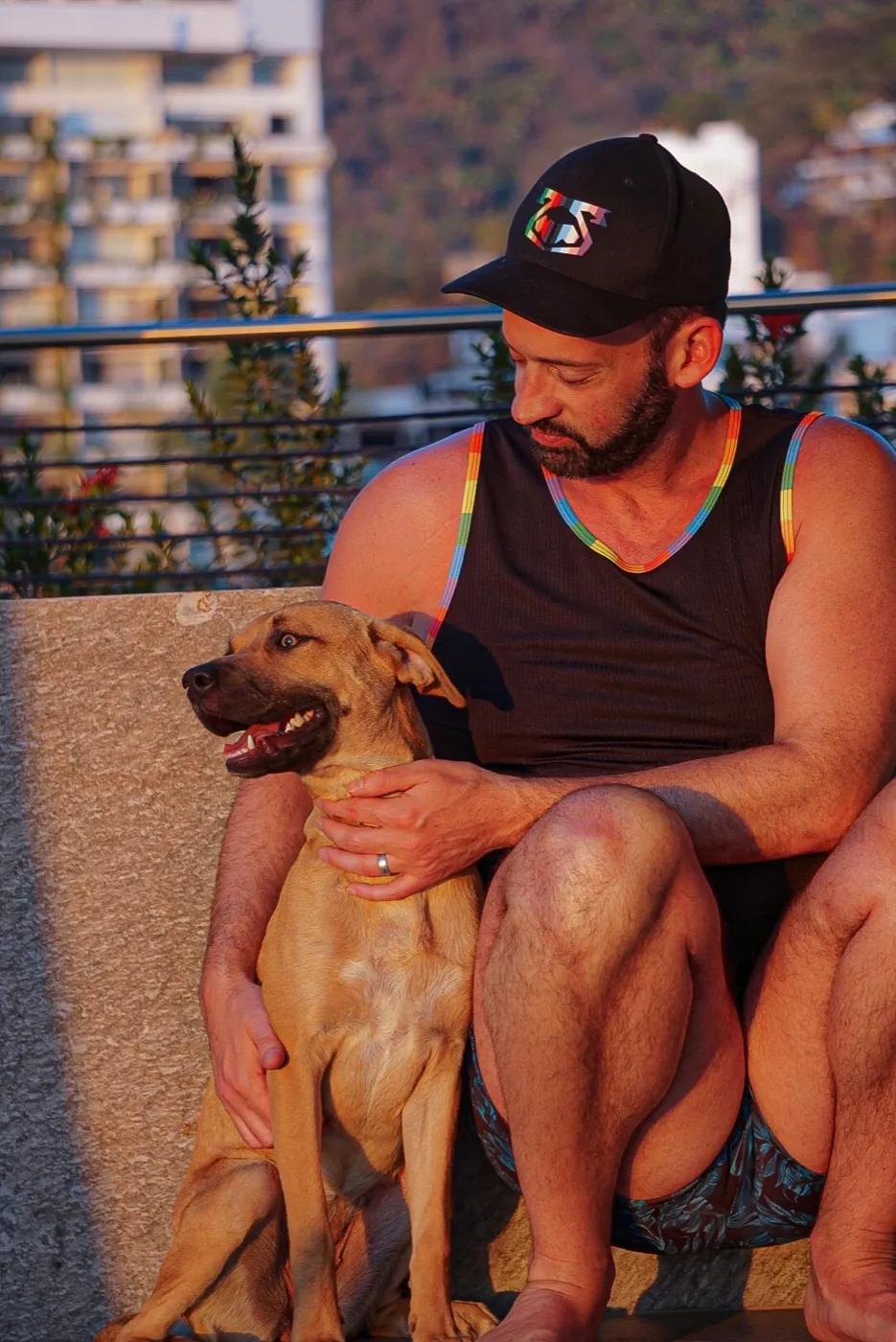 A man in a cap sits with a relaxed dog at golden hour above a hillside town